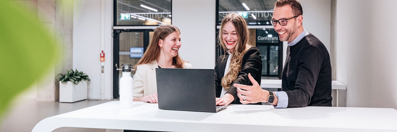 Drei Mitarbeitende lachen gemeinsam bei einer Besprechung an einem modernen Arbeitsplatz. Das Bild steht für gelebte Teamkultur und die attraktiven Benefits, die bei Daimler Truck zu einem motivierenden Arbeitsumfeld beitragen.