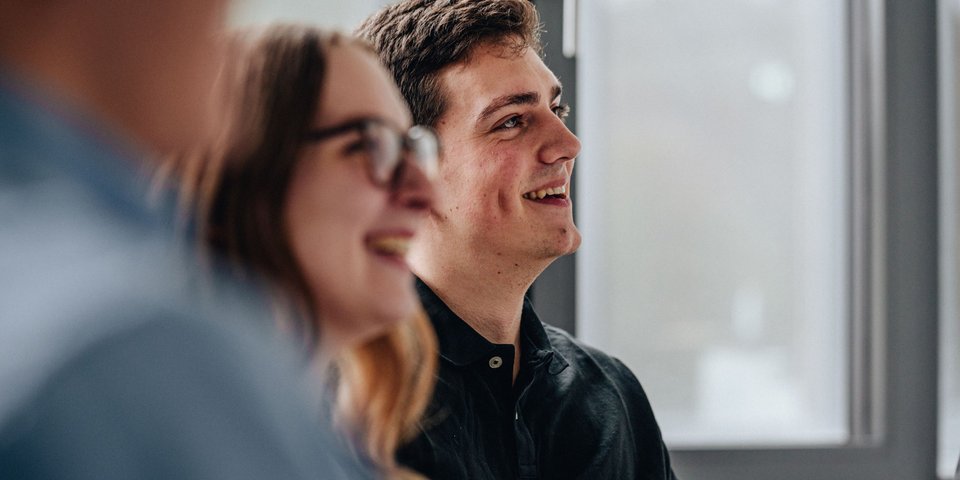 Zwei junge duale Studierende lächeln, während sie in einem lichtdurchfluteten Raum an einem Gespräch teilnehmen.