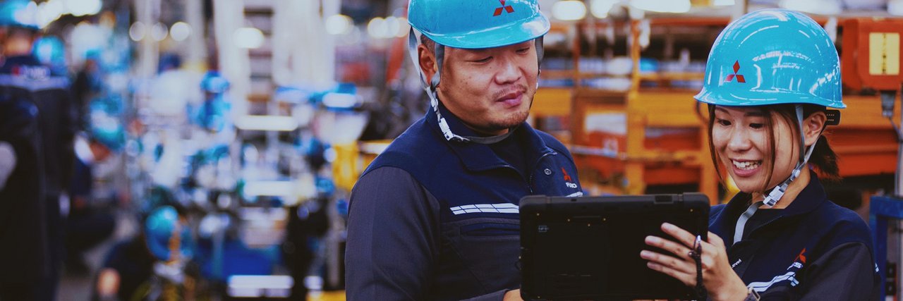 Two production employees wearing blue hard hats look at a process monitoring tablet in a busy production hall.