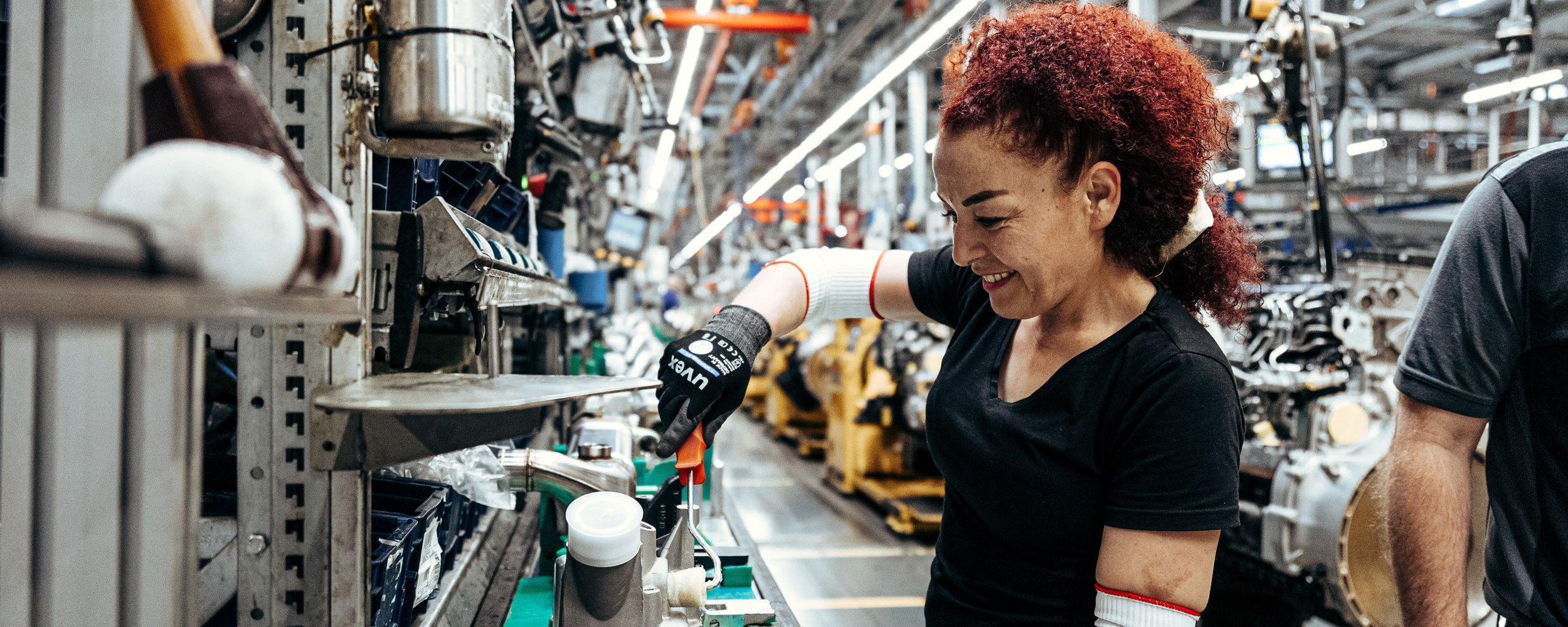 A red-haired woman wearing safety gloves works on a Daimler Truck production part in a production hall.