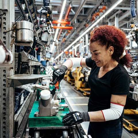Eine rothaarige Frau arbeitet in einer Produktionshalle mit Sicherheitshandschuhen an einem Daimler Truck Fertigungsteil.
