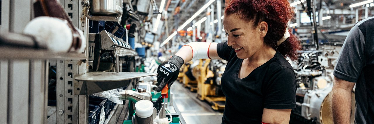 Eine rothaarige Frau arbeitet in einer Produktionshalle mit Sicherheitshandschuhen an einem Daimler Truck Fertigungsteil.