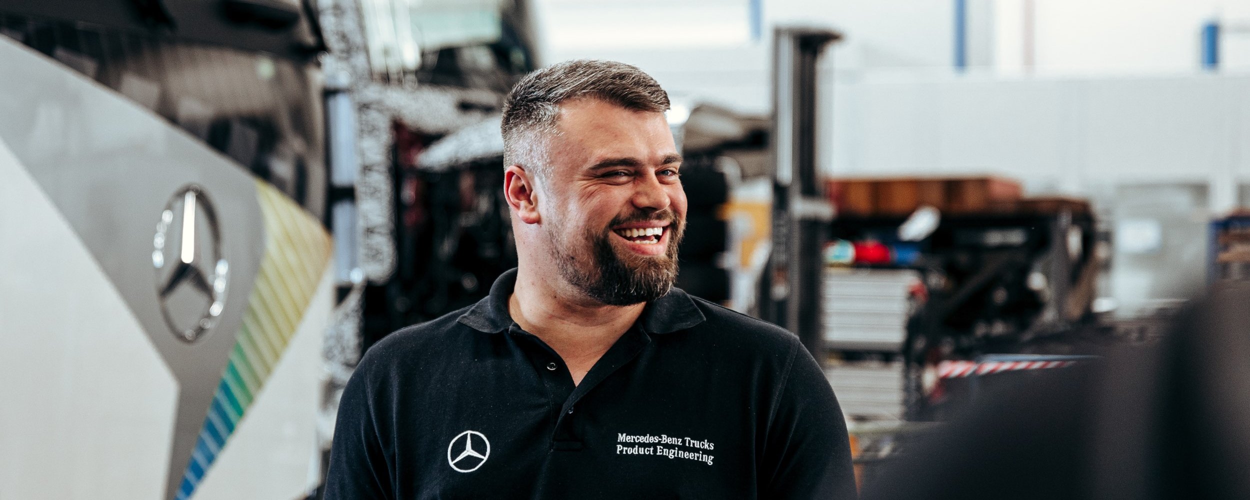 Mitarbeiter in schwarzem Poloshirt lächelt in einer Werkstatt, im Hintergrund ist ein Daimler Truck Lkw zu sehen.