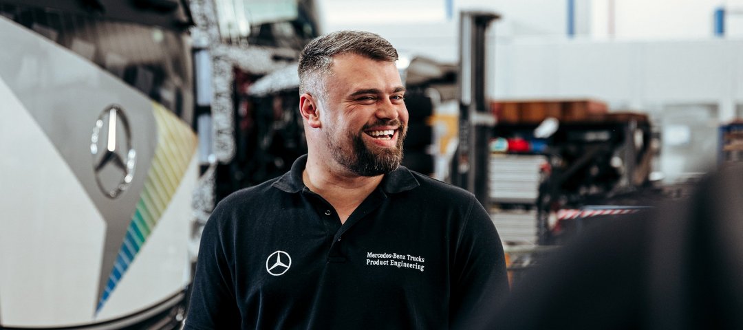 Mitarbeiter in schwarzem Poloshirt lächelt in einer Werkstatt, im Hintergrund ist ein Daimler Truck Lkw zu sehen.
