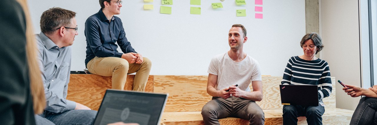 Mitarbeitende diskutieren in einem modern gestalteten Arbeitsraum vor einer strukturierten Post-it-Wand. Das Bild steht für kreative Arbeitsprozesse und agile Methoden im Alltag bei Daimler Truck.