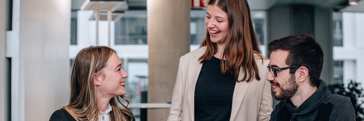 Lachende junge Absolventen in einer Teamdiskussion, während sie gemeinsam an dem Laptop vor ihnen arbeiten.