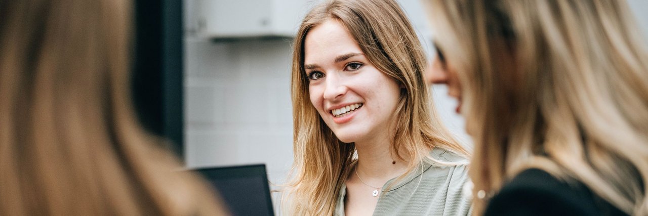 Eine junge Frau lächelt und schaut in die Runde, während sie mit Kolleginnen an einem Tisch mit einem Laptop sitzt.