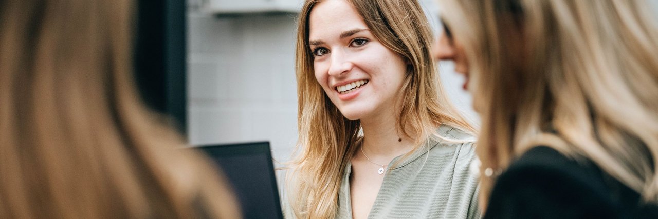 A young woman smiles and looks round as she sits with colleagues at a table with a laptop.