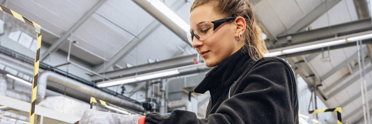 Eine technische Auszubildende mit Schutzbrille und Sicherheitshandschuhen arbeitet an einem Schraubstock in einer Werkstattumgebung.