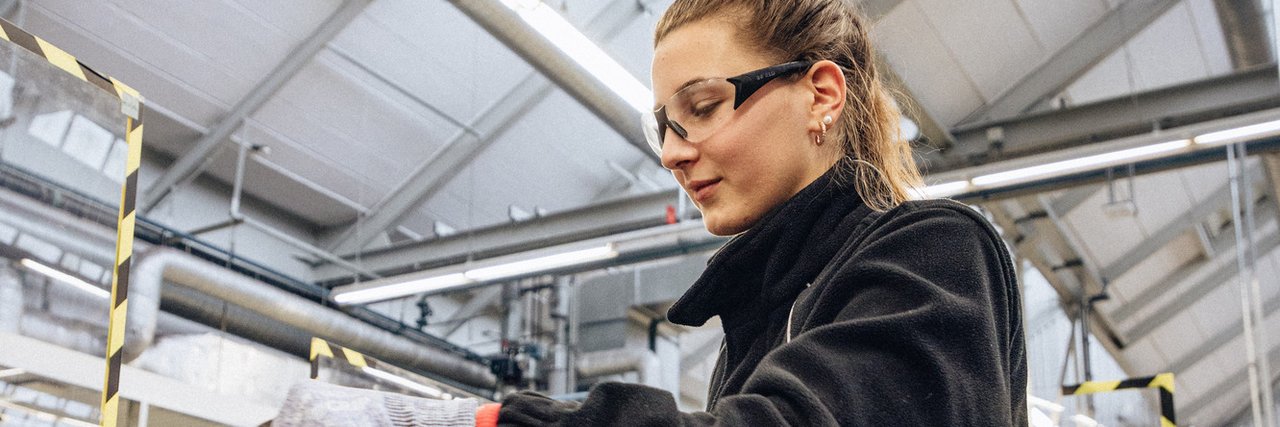 Eine technische Auszubildende mit Schutzbrille und Sicherheitshandschuhen arbeitet an einem Schraubstock in einer Werkstattumgebung.