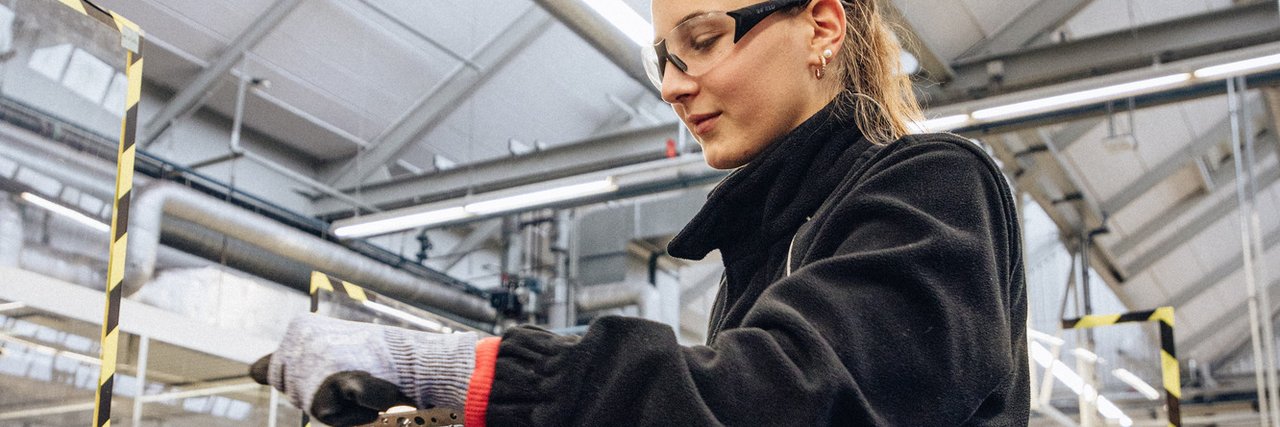 A technical apprentice wearing safety goggles and safety gloves works on a vice in a workshop environment.