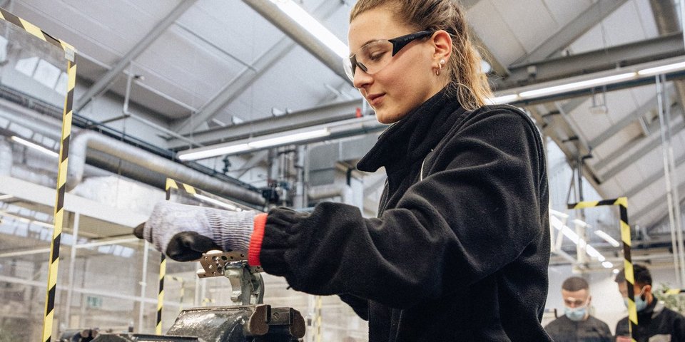 Eine technische Auszubildende mit Schutzbrille und Sicherheitshandschuhen arbeitet an einem Schraubstock in einer Werkstattumgebung.