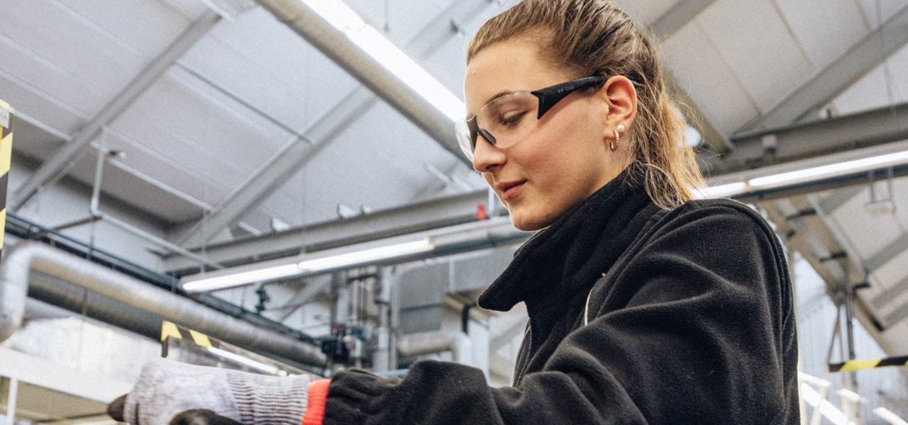 Eine technische Auszubildende mit Schutzbrille und Sicherheitshandschuhen arbeitet an einem Schraubstock in einer Werkstattumgebung.
