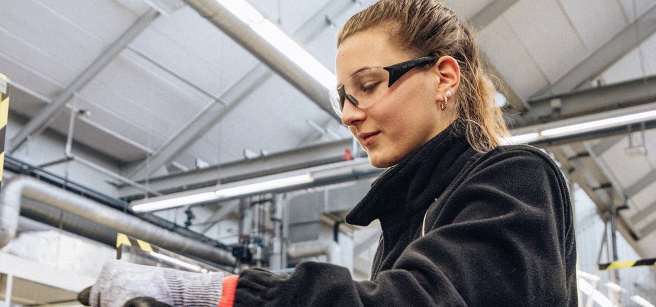 Eine technische Auszubildende mit Schutzbrille und Sicherheitshandschuhen arbeitet an einem Schraubstock in einer Werkstattumgebung.