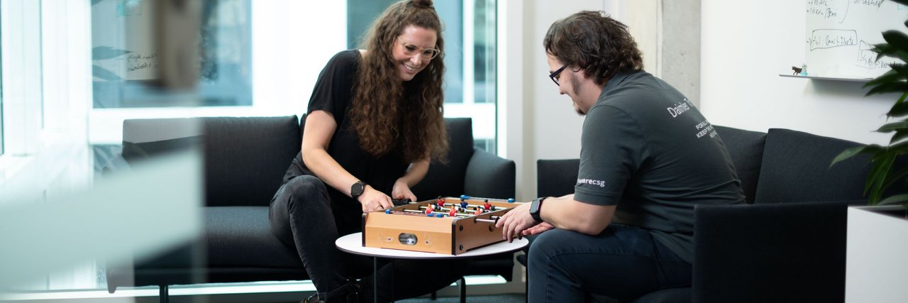 Two employees sit on a sofa in the Daimler Truck Campus and play mini table football together.