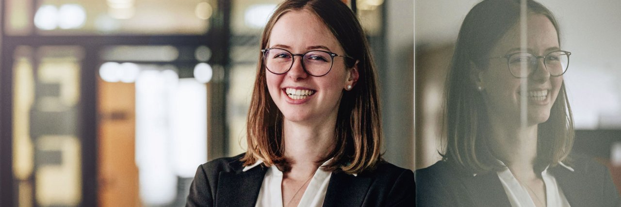 A young Daimler truck employee in a business outfit stands smiling confidently in front of a glass wall. The picture symbolises professional success and the recognition that comes with fair and attractive compensation.