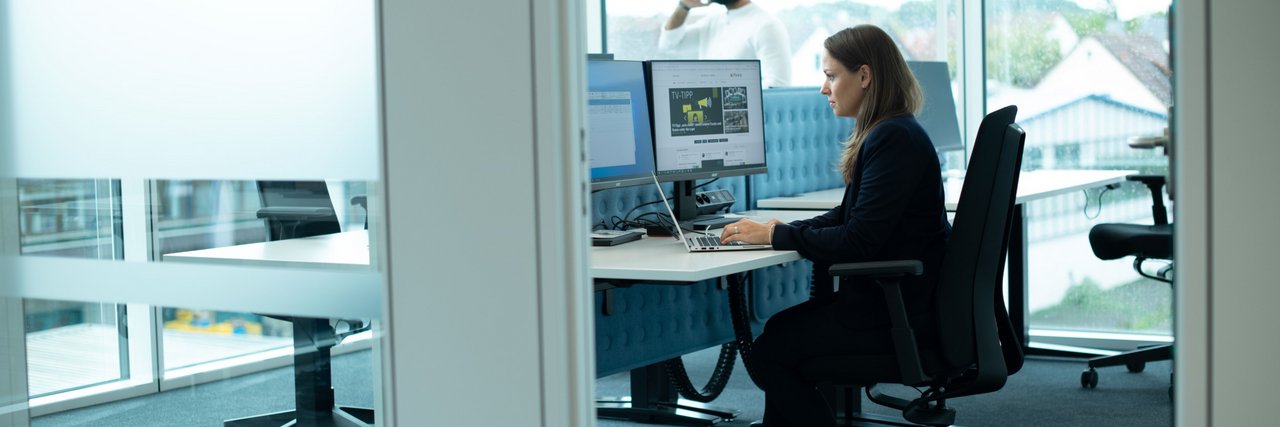 A woman in business attire works intently at two monitors in the Legal & Compliance department of Daimler Truck. A man is on the phone in the background.