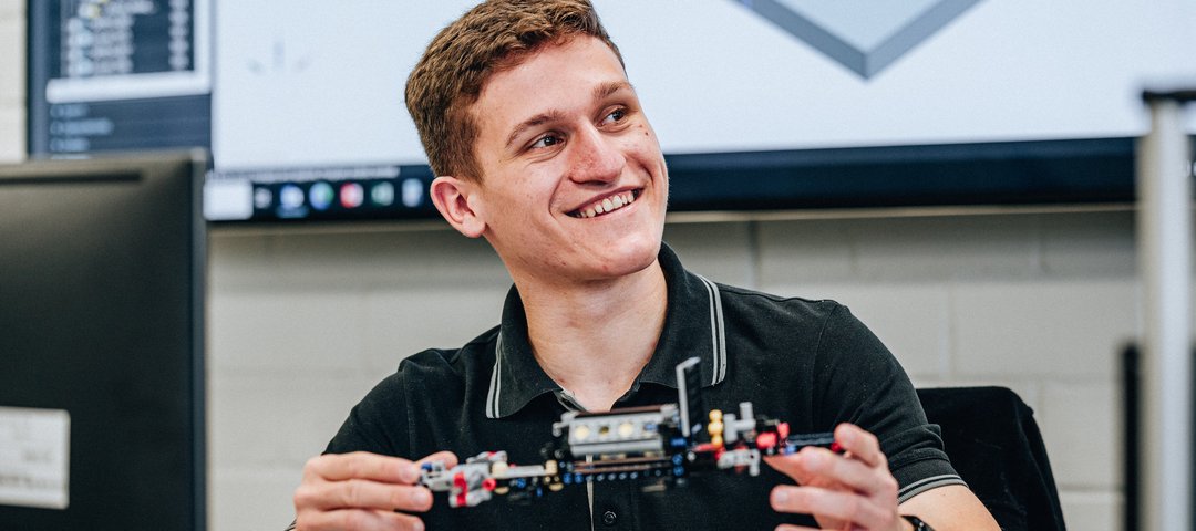 A dual student smiles as he shows a technical model made of building blocks, with a monitor with CAD software in the background.