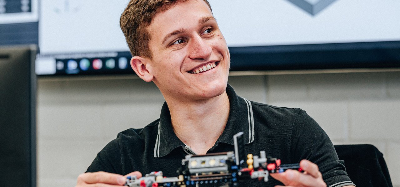 Ein dualer Student zeigt lächelnd ein technisches Modell aus Bausteinen, im Hintergrund ein Monitor mit CAD-Software.