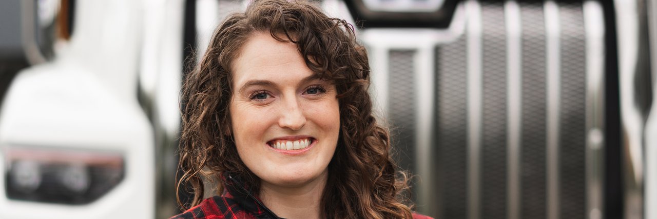 Portrait of Alex Banzer, a smiling woman with curly hair and folded arms stands in front of a large white truck.