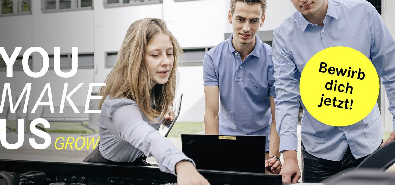 Eine Studentin zeigt auf eine technische Komponente an einem Truck und hält ein Tablet, während die anderen zwei Studenten aufmerksam zuschauen. Der Slogan 'You make us grow' ist auf der linken Seite zu sehen, begleitet von einem gelben Button mit 'Bewirb dich jetzt!'