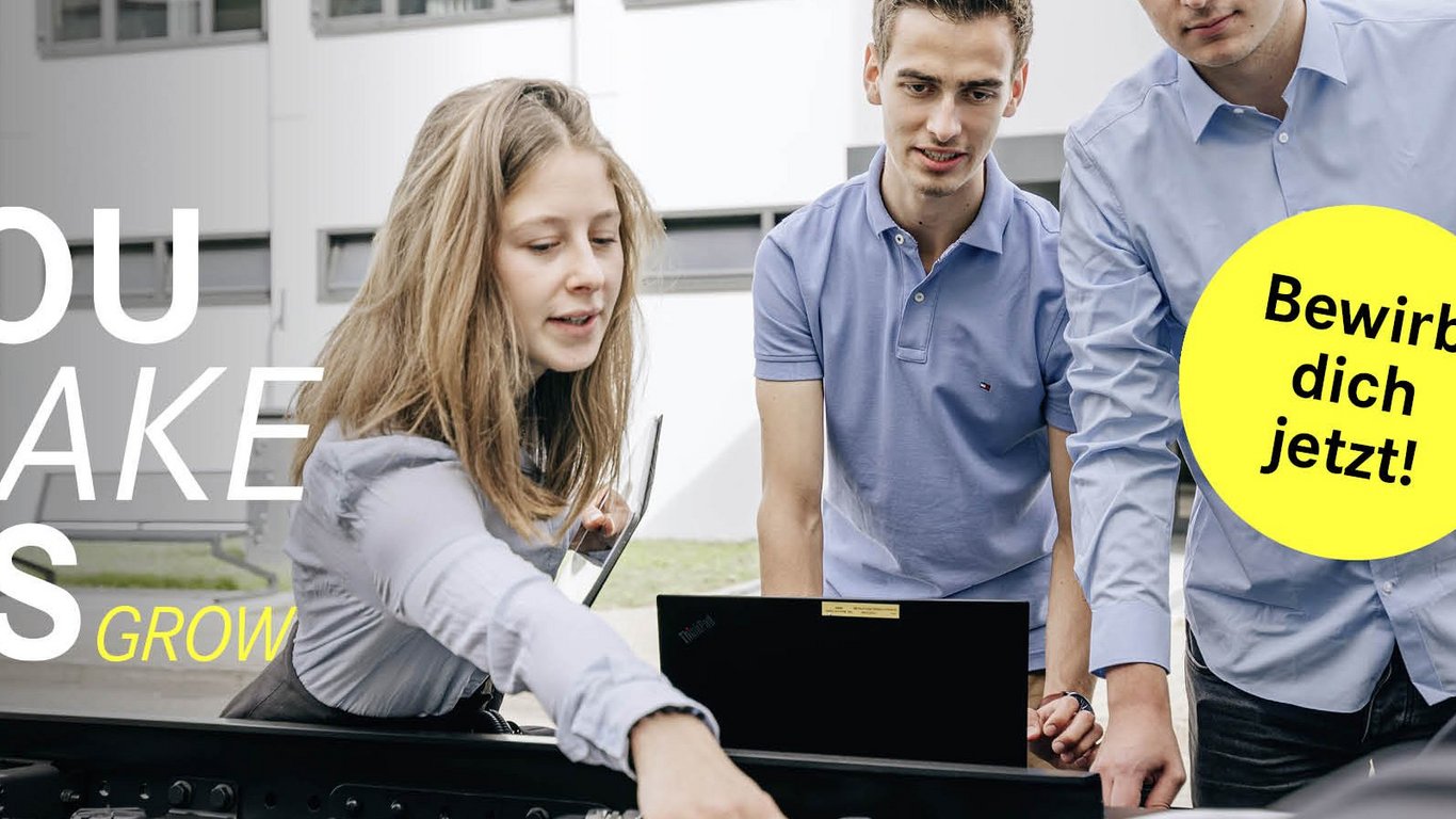 Eine Studentin zeigt auf eine technische Komponente an einem Truck und hält ein Tablet, während die anderen zwei Studenten aufmerksam zuschauen. Der Slogan 'You make us grow' ist auf der linken Seite zu sehen, begleitet von einem gelben Button mit 'Bewirb dich jetzt!'