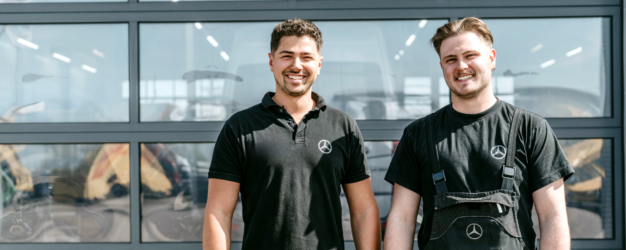 Two technicians from Daimler Truck's commercial vehicle centers stand smiling in front of a modern workshop building. Both are wearing black work clothes with the Mercedes-Benz logo.