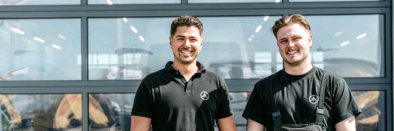 Zwei Techniker der Nutzfahrzeugzentren von Daimler Truck stehen lächelnd vor einer modernen Werkstatthalle. Beide tragen schwarze Arbeitskleidung mit Mercedes-Benz Logo.