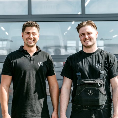 Two technicians from Daimler Truck's commercial vehicle centers stand smiling in front of a modern workshop building. Both are wearing black work clothes with the Mercedes-Benz logo.