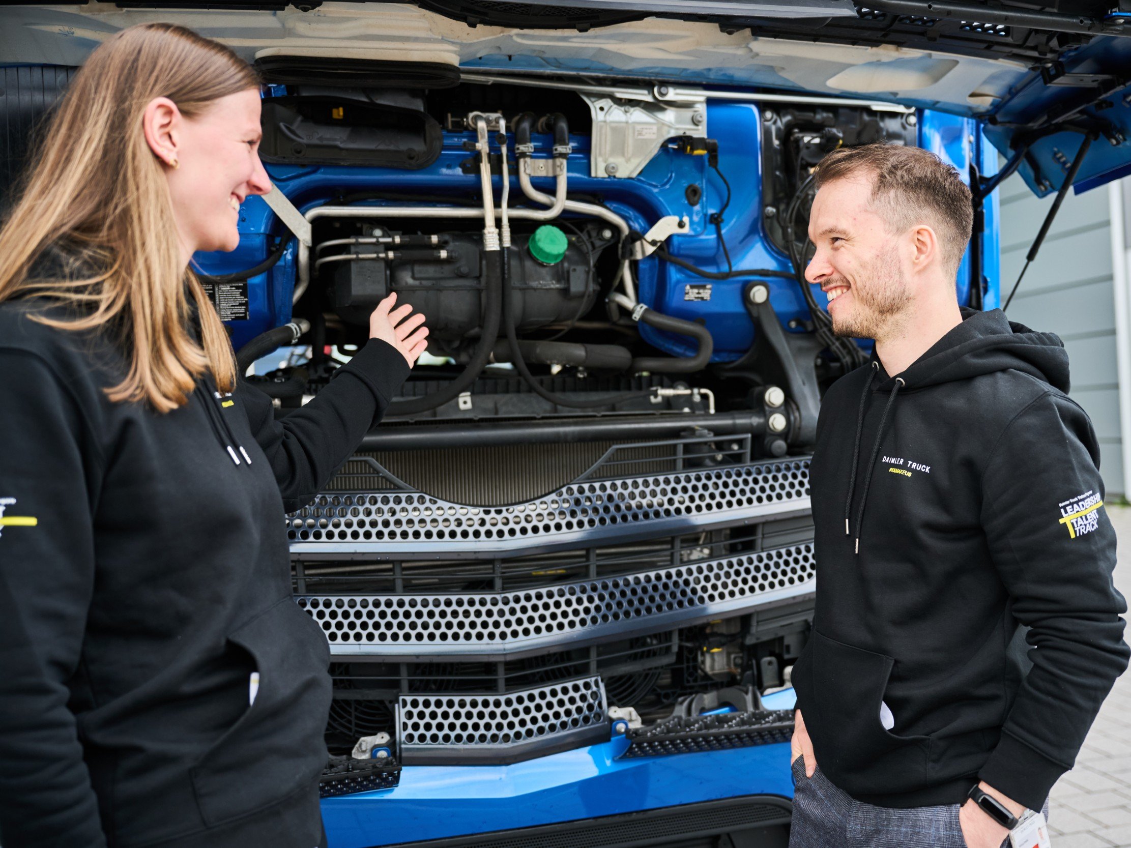 Zwei Teilnehmende des Daimler Truck Leadership Talent Track Traineeprogramms stehen vor einem geöffneten Lkw-Motor und diskutieren technische Details – Teil des Moduls „Customer First @ First Retail“.