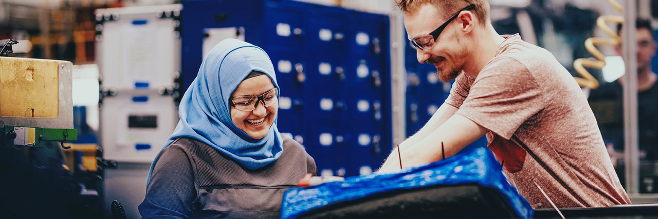 A woman with a headscarf and a man with safety goggles are smiling as they work together on a component in an assembly hall.