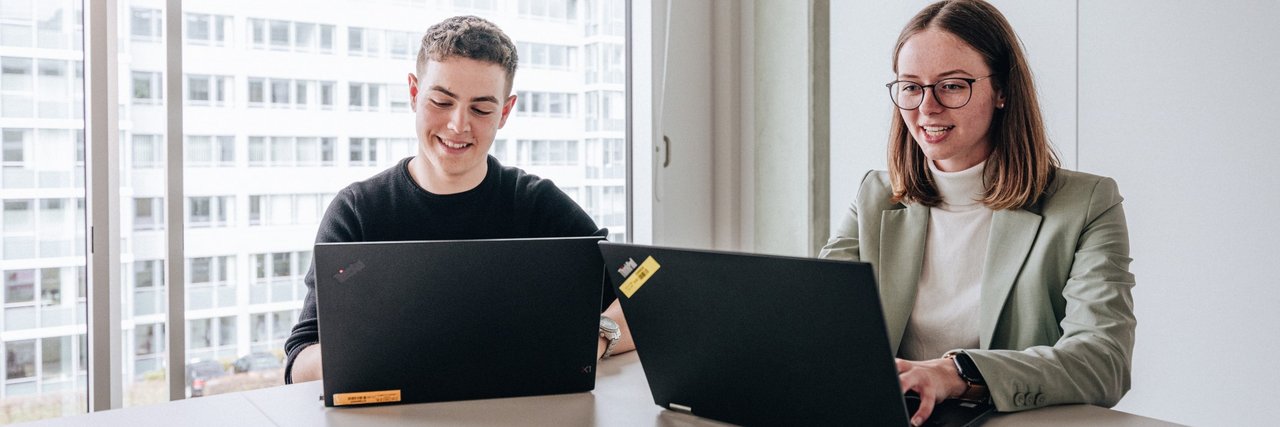 Zwei Doktoranten, ein junger Mann und eine junge Frau, sitzen nebeneinander und arbeiten am Laptop im Daimler Truck Campus.