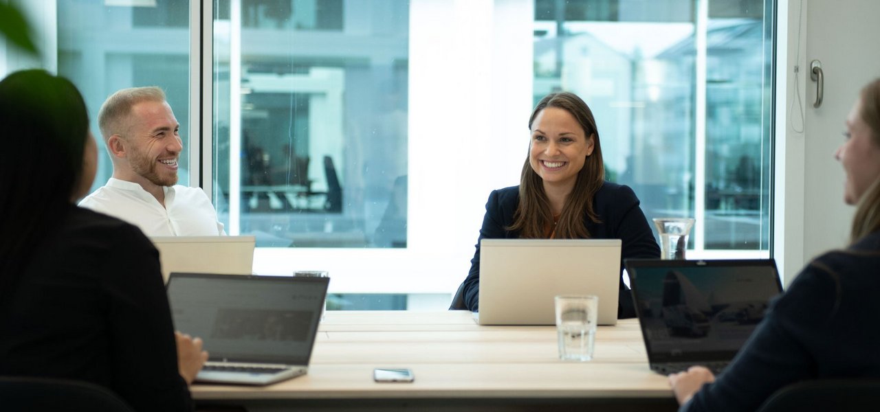 Eine Besprechung in einem modernen Büro, bei der sich vier Mitarbeitende entspannt unterhalten und Notizen auf ihren Laptops machen.