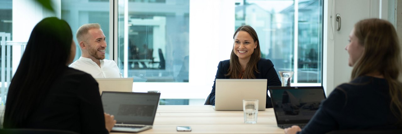 A meeting in a modern office where four employees are chatting in a relaxed atmosphere and taking notes on their laptops.