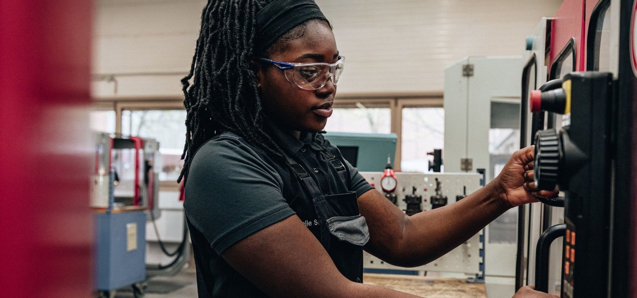Eine Mitarbeiterin in Arbeitskleidung und mit Schutzbrille bedient konzentriert eine Maschine in einer Werkshalle.