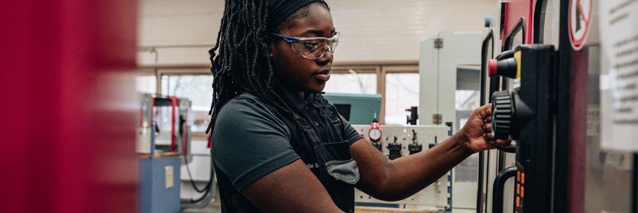 Eine Mitarbeiterin in Arbeitskleidung und mit Schutzbrille bedient konzentriert eine Maschine in einer Werkshalle.