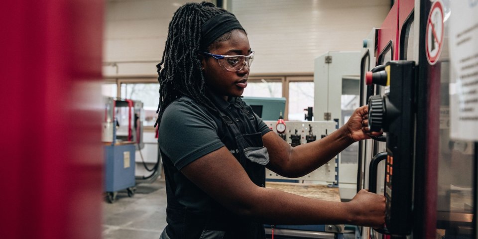 Eine Mitarbeiterin in Arbeitskleidung und mit Schutzbrille bedient konzentriert eine Maschine in einer Werkshalle.