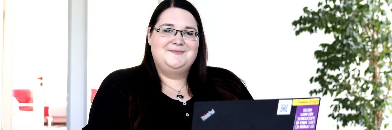 Diana Andrä sits smiling at a desk with a laptop in a bright, modern environment.