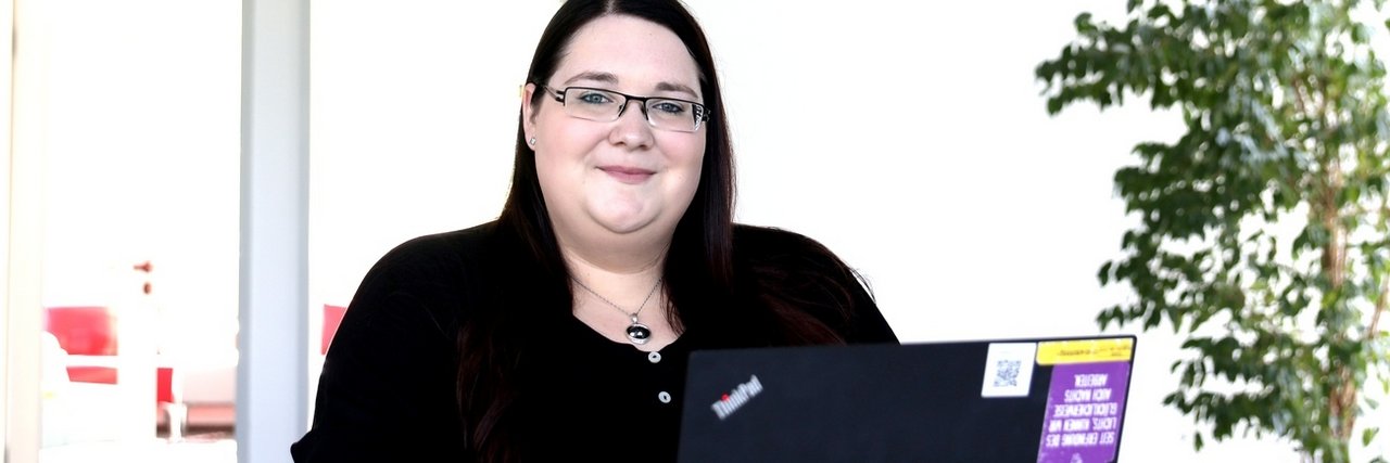 Diana Andrä sitzt lächelnd an einem Schreibtisch mit einem Laptop in einer hellen, modernen Umgebung.