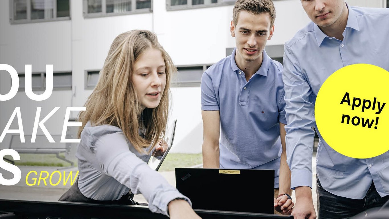 Eine Studentin zeigt auf eine technische Komponente an einem Truck und hält ein Tablet, während die anderen zwei Studenten aufmerksam zuschauen. Der Slogan 'You make us grow' ist auf der linken Seite zu sehen, begleitet von einem gelben Button mit 'Apply now!'