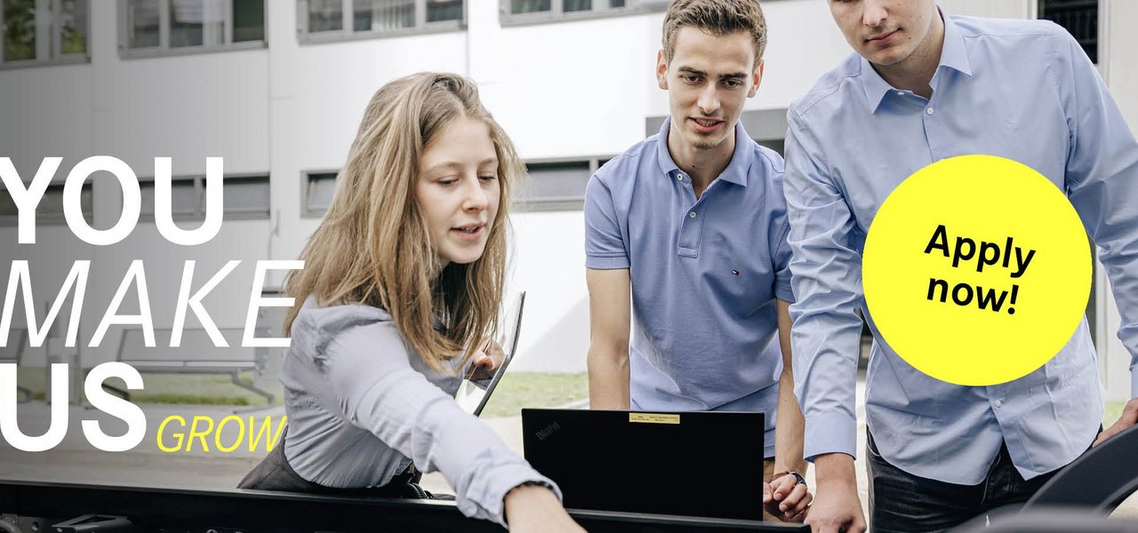 A female student points to a technical component on a truck and holds a tablet while the other two students look on attentively. The slogan "You make us grow" can be seen on the left-hand side, accompanied by a yellow button with "Apply now!"
