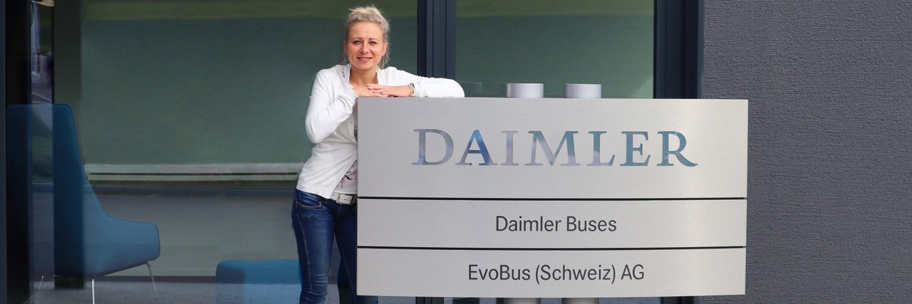 Die PR-Managerin von Daimler Buses Aylin Flegel lehnt an einem Firmenschild mit dem Daimler-Logo vor einem modernen Bürogebäude.