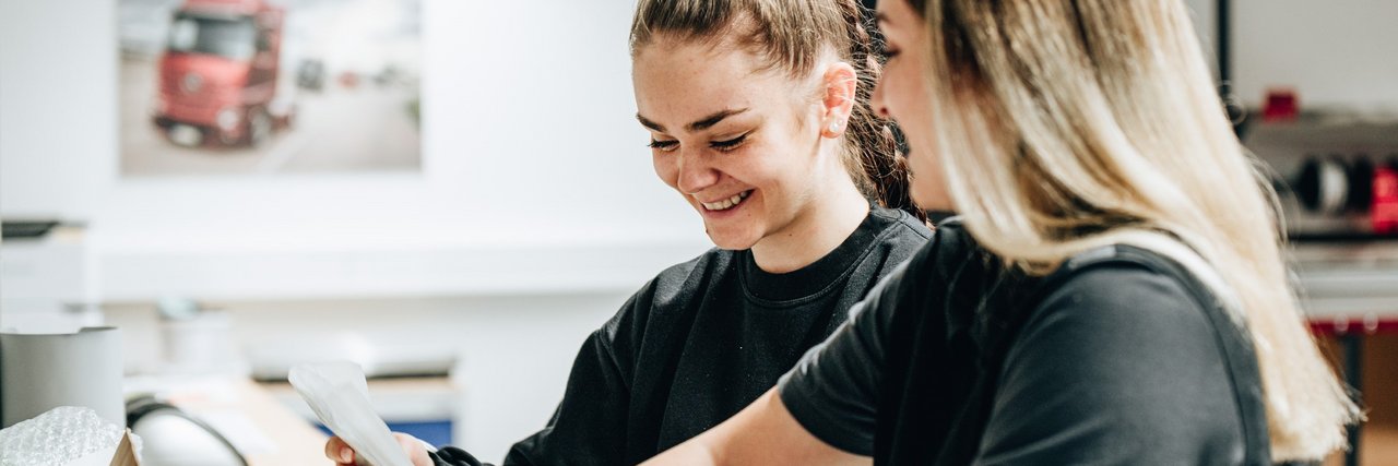 Zwei fröhliche Auszubildende in schwarzer Arbeitskleidung verpacken gemeinsam eine Kiste.