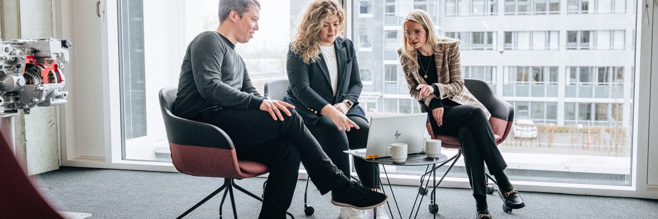 Three people are sitting in a brightly lit room on the Daimler Truck Campus, discussing something on a laptop placed on a small table.