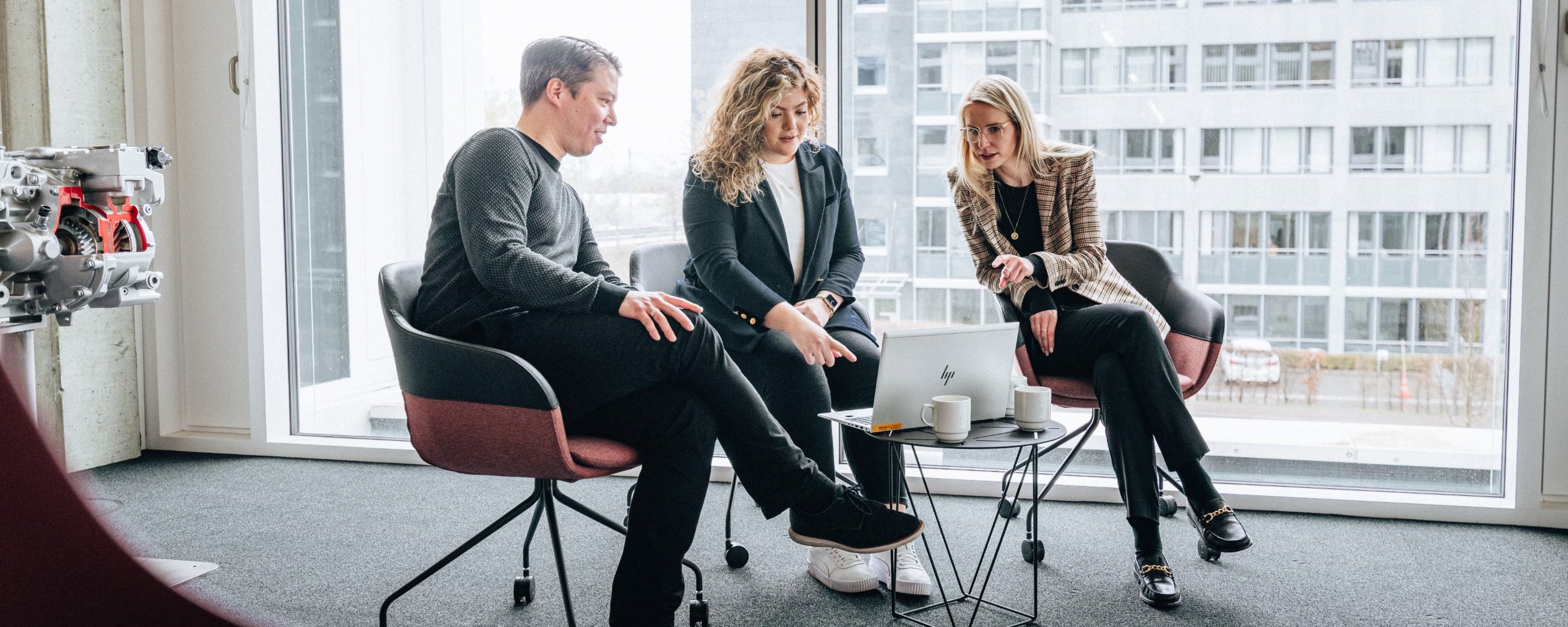 Drei Personen sitzen in einem hellen Raum auf dem Daimler Truck Campus und besprechen etwas an einem Laptop, der auf einem kleinen Tisch steht.