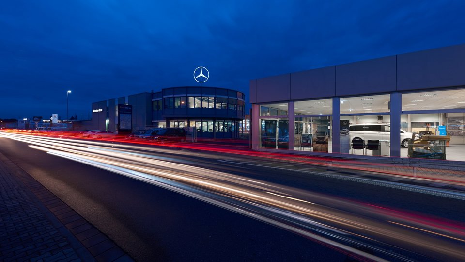 Mercedes-Benz Nutzfahrzeugzentrum Weiterstadt bei Nacht, mit beleuchteten Ausstellungsräumen und Lichtspuren vorbeifahrender Fahrzeuge.