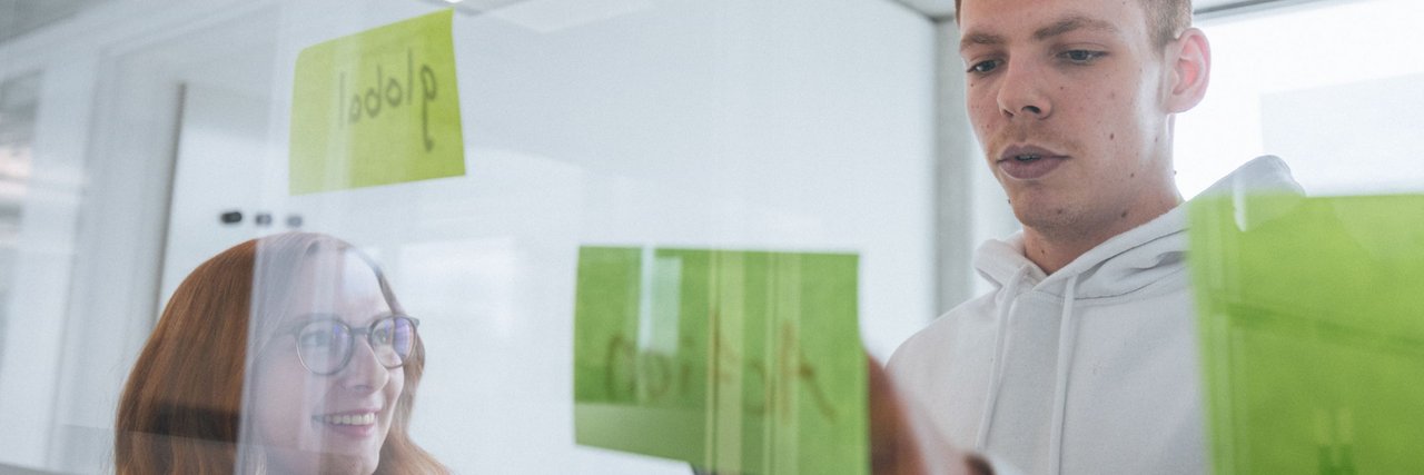 Two young employees stand behind a glass wall with colourful sticky notes, the woman smiles while the man arranges a note.