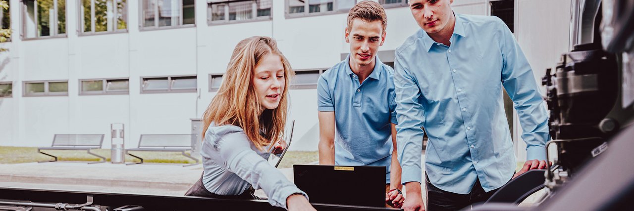 Three young students are working together on a vehicle frame in the open air. One student points to a technical component and holds a tablet while the other two students watch attentively.