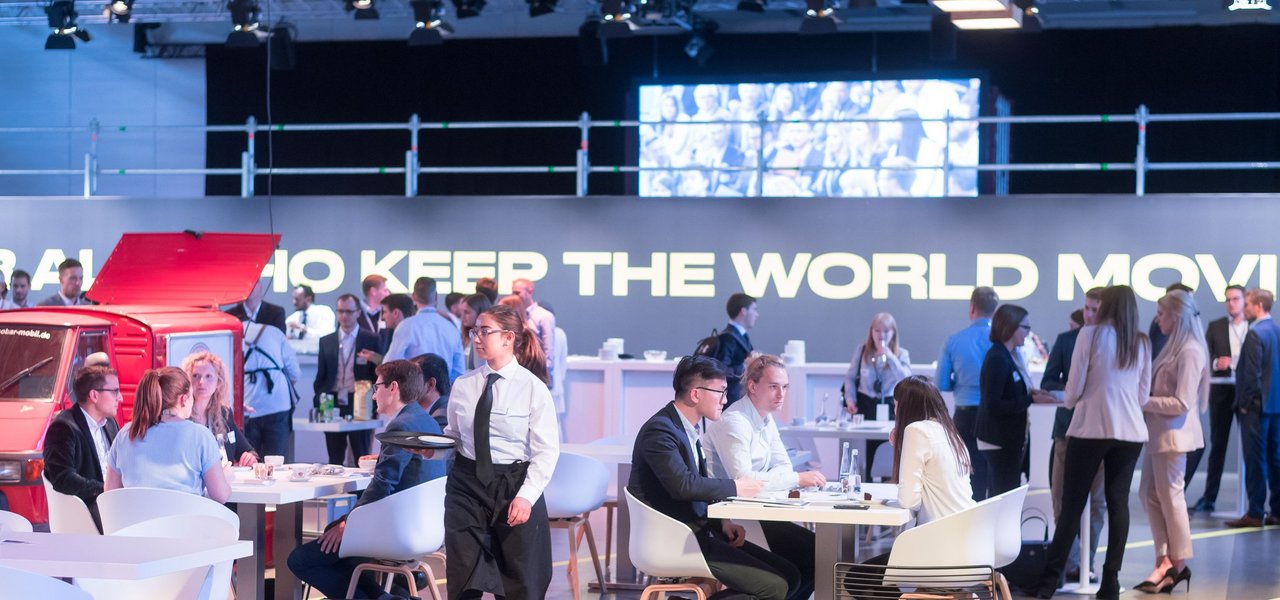 Karriere Event von Daimler Truck im eleganten Business Setting mit Gästen im Austausch und einem großen Schriftzug „FOR ALL WHO KEEP THE WORLD MOVING“ im Hintergrund.