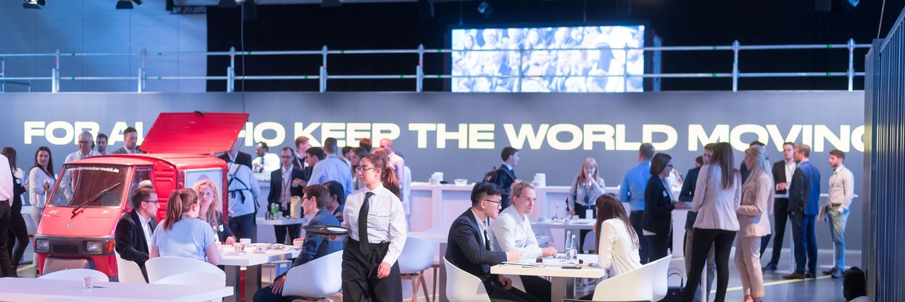 Karriere Event von Daimler Truck im eleganten Business Setting mit Gästen im Austausch und einem großen Schriftzug „FOR ALL WHO KEEP THE WORLD MOVING“ im Hintergrund.